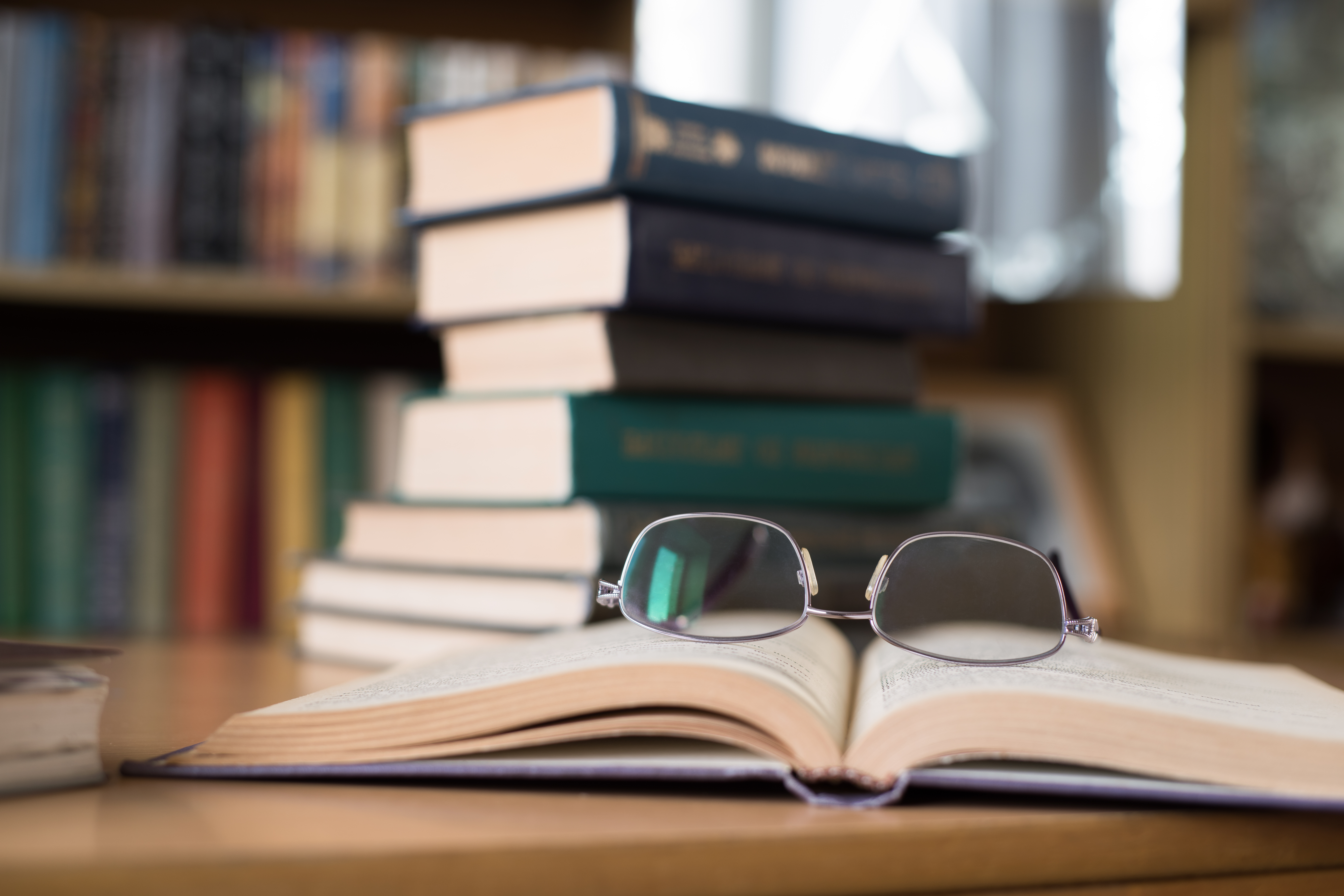 Opened Book with Glasses above and a Stack of Books on the Background